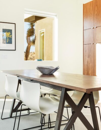 Modern kitchen interior with a wooden table, white chairs, and a view of a desert landscape through large windows.