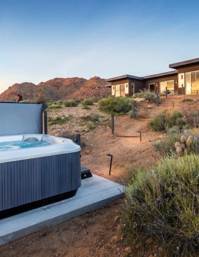 A hot tub with a cover partially lifted, in front of a modern home in a desert landscape at twilight.