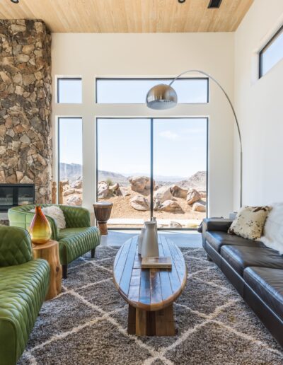 Modern living room with large windows showcasing desert views, featuring a stone fireplace, leather sofas, and an arching floor lamp.