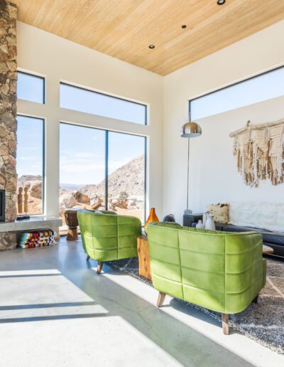 Modern living room with high ceilings, large windows, stone fireplace, and green accent chairs.