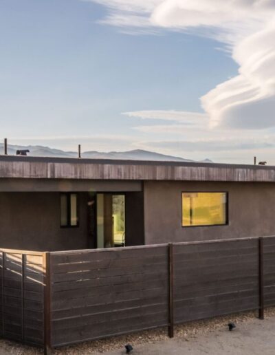 Modern single-story home with a flat roof and large windows, featuring a dark wood fence, under a vast sky with distinctive cloud formations.