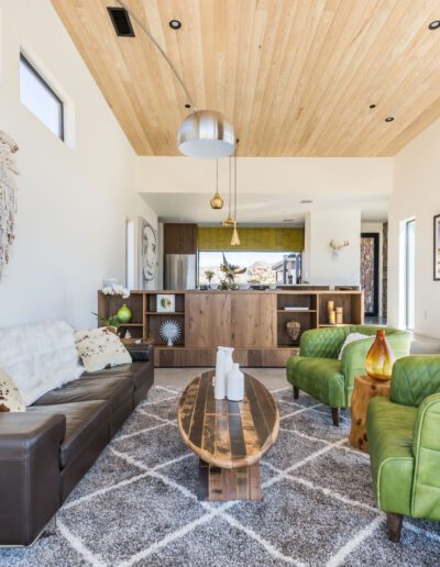 Modern living room interior featuring a brown leather sofa, a green armchair, wooden ceiling, and decorative elements, with an adjoining open kitchen in the background.