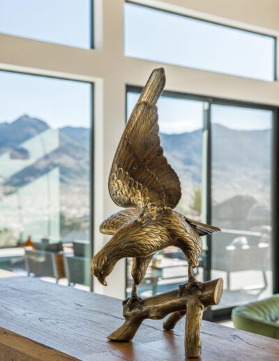 Bronze eagle sculpture displayed on a wooden surface with large windows overlooking a mountainous landscape in the background.