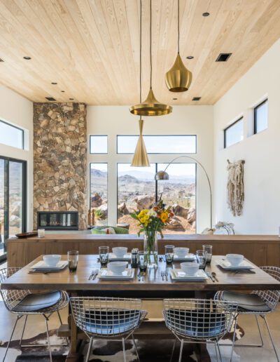 Modern dining room with natural light, a wooden ceiling, and a stone accent wall, overlooking a desert landscape.