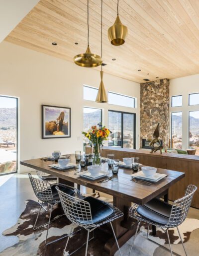 Modern dining room with large windows offering a desert view, featuring a wooden table, stylish chairs, and pendant lights.