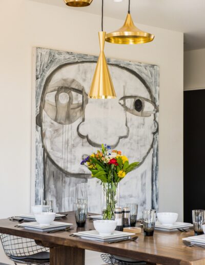 Elegant dining table set for a meal beneath modern pendant lights, with a large abstract face painting as the backdrop.