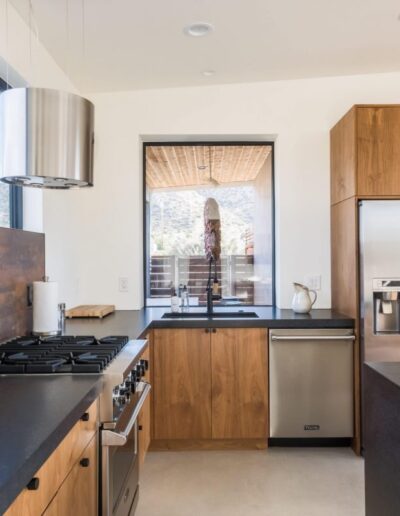 Modern kitchen with wooden cabinets, stainless steel appliances, and a central island with a cooktop and hood, overlooking a mountainous landscape through a large window.