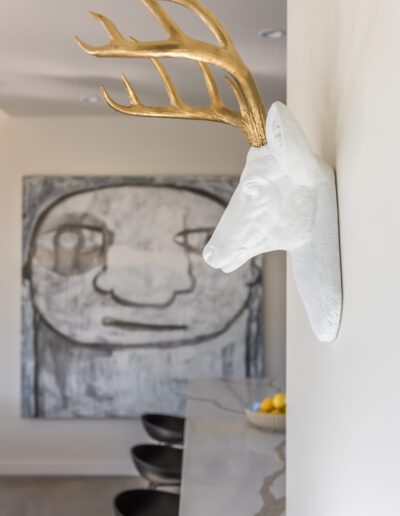 A white deer head sculpture with golden antlers mounted on a wall in a modern interior alongside a large monochromatic artwork and a kitchen counter with stools.