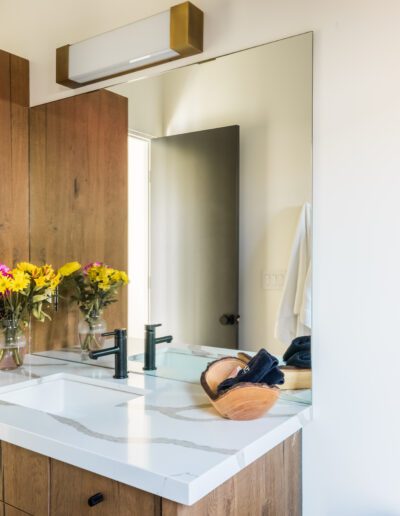 A modern bathroom with wooden cabinets, a white countertop with a sink, a mirror, and fresh flowers, with a framed owl print on the wall.