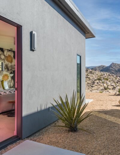 A modern desert home with an open sliding door revealing a cozy bedroom, surrounded by arid landscape and cacti.