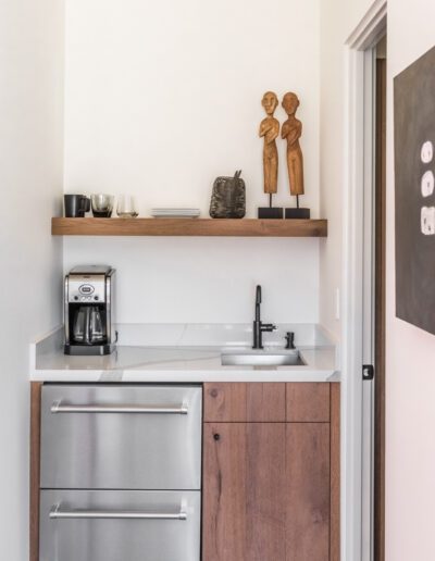 A modern kitchenette with wooden cabinets, a coffee maker, and decorative items on a shelf.