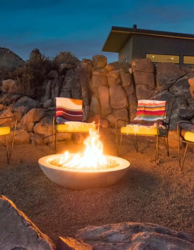 Outdoor seating area with a fire pit and colorful blankets on chairs at dusk, with a crescent moon in the sky.