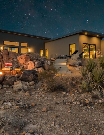 Modern desert home at twilight with lit interior and outdoor fire pit under a starry sky.