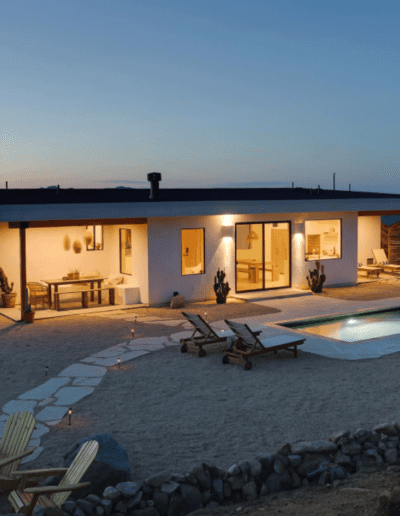 Modern single-story house with illuminated interior and exterior, featuring a swimming pool and outdoor seating area, during twilight.