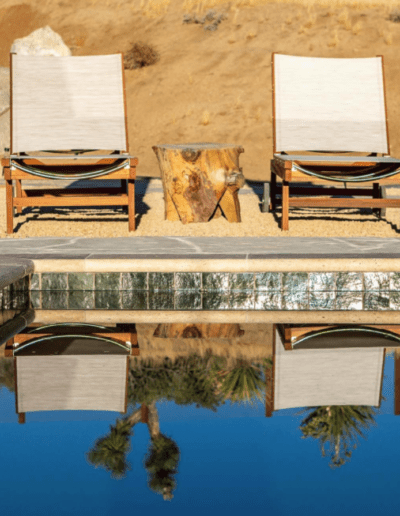 Two lounge chairs by a pool with their reflection on the water's surface.