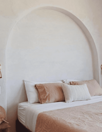 A minimalist bedroom featuring a neatly made bed with beige linens, flanked by two wall-mounted lamps, against a textured off-white wall with a large arch-shaped niche.