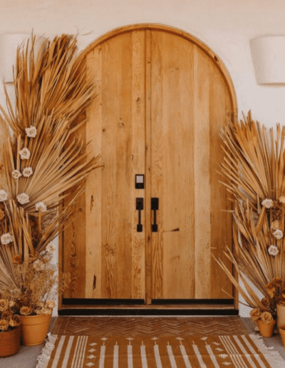 Arched wooden door flanked by decorative dried grass arrangements and potted plants.