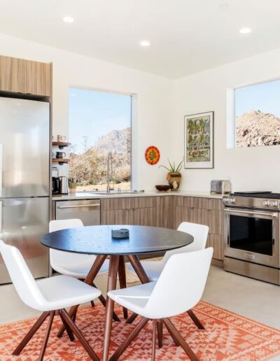 Modern kitchen with stainless steel appliances and a round dining table with white chairs, set against a backdrop of mountain views through large windows.