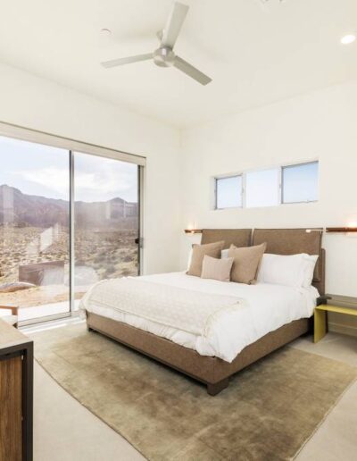 A modern bedroom with large windows offering a desert view, featuring a queen-sized bed, ceiling fan, and minimalistic decor.