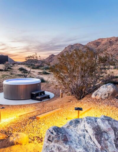 A modern outdoor hot tub with a cover set against a desert landscape at sunset.