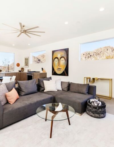 A modern living room with a gray sectional sofa, a glass coffee table, decorative artwork, and a desert landscape visible through the windows.